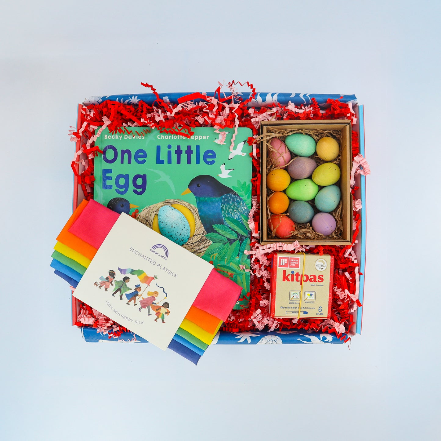 A brightly coloured children’s gift box containing colourful wooden eggs, a rainbow playsilk, a packet of kitpas crayons and the picture book One Little Egg by Becky Davies.