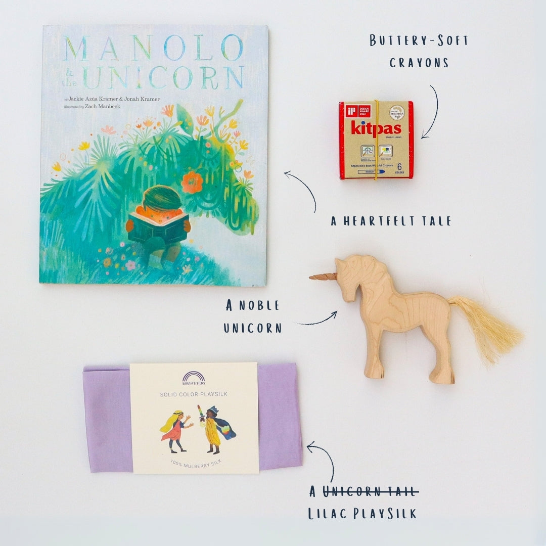 A display of The Unicorn StoryBox contents including a wooden unicorn, a lilac playsilk, a packet of kitpas crayons and the picture book Manolo and the Unicorn.