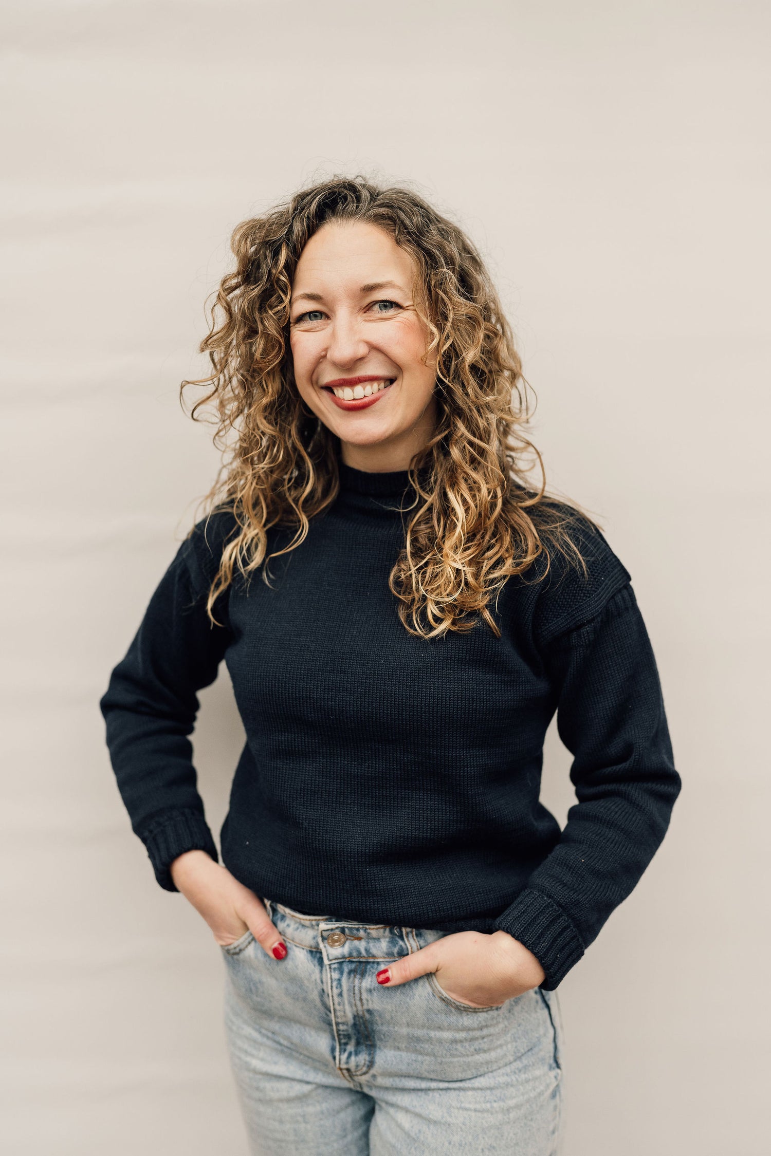 Picture of a smiling woman with curly hair, wearing jeans and a navy jumper.