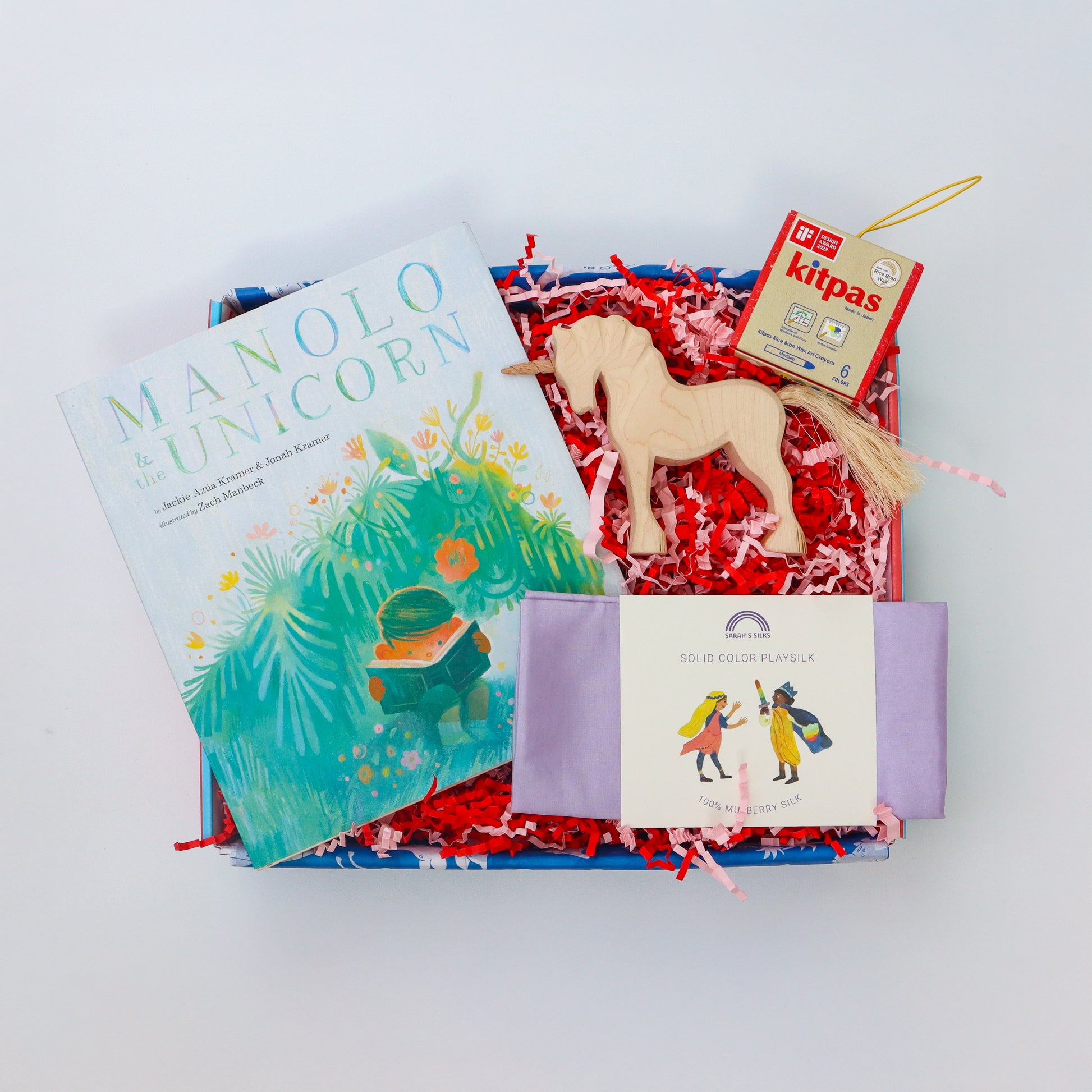 A brightly coloured children’s gift box containing a wooden unicorn, a lilac playsilk, a packet of kitpas crayons and the picture book Manolo and the Unicorn.