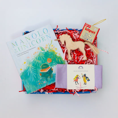A brightly coloured children’s gift box containing a wooden unicorn, a lilac playsilk, a packet of kitpas crayons and the picture book Manolo and the Unicorn.